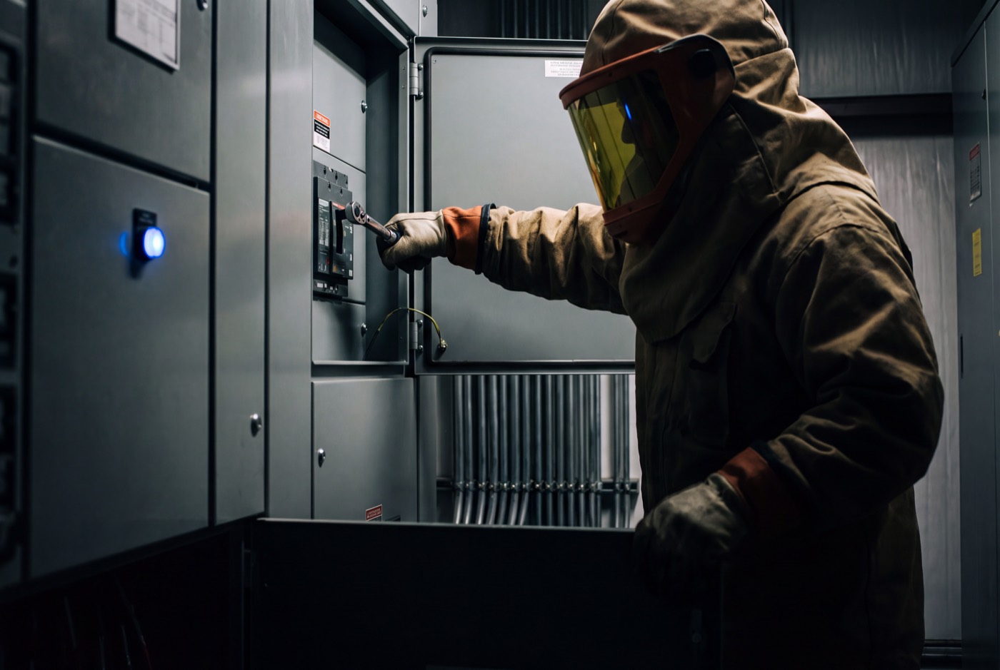 A worker in a full arc-flash suit and hood turning a breaker with a wrench inside an open switchgear cabinet
